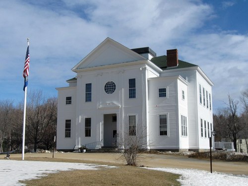 A white historic building with a large circular window, surrounded by snow and grass, under a cloudy sky.