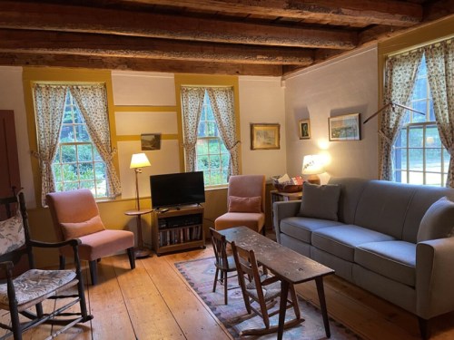 Cozy living room with wooden beams, two windows, a sofa, armchairs, a TV, and a small table with chairs.