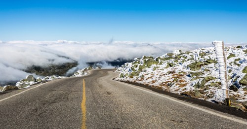 A winding road through a snowy landscape, with clouds below and a clear blue sky above.