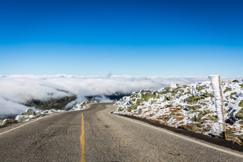 A winding road through a snowy landscape, with clouds below and a clear blue sky above.