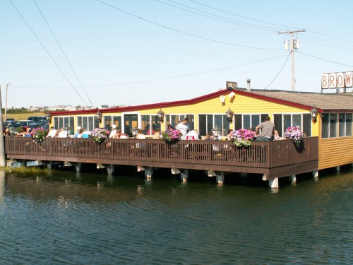 A yellow waterfront restaurant with outdoor seating, surrounded by water and colorful flower pots.