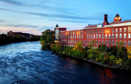 A brick building along a river at sunset, with trees and a clear sky reflecting on the water's surface.