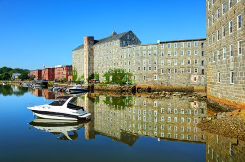 A boat floats on calm water, reflecting historic stone buildings along the shore under a clear blue sky.