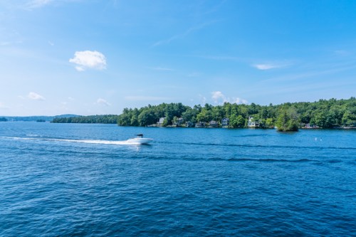 A boat glides across a blue lake, surrounded by lush green trees and distant houses under a clear sky.