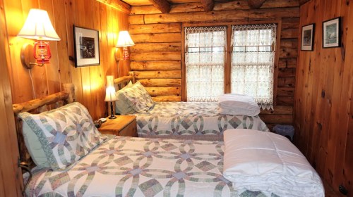 Cozy log cabin bedroom with two twin beds, quilted bedding, wooden walls, and warm lighting.