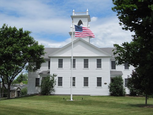 A white building with a bell tower, featuring an American flag on a pole in front, surrounded by green trees and grass.