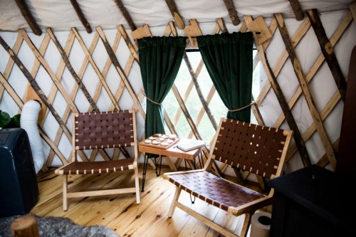 Cozy interior of a yurt featuring two wooden chairs, a small table, and green curtains against a lattice wall.