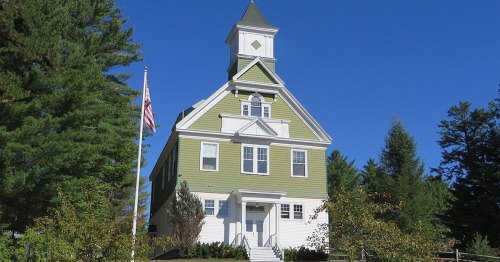 A tall, green Victorian-style house with a clock tower, surrounded by trees and an American flag in front.