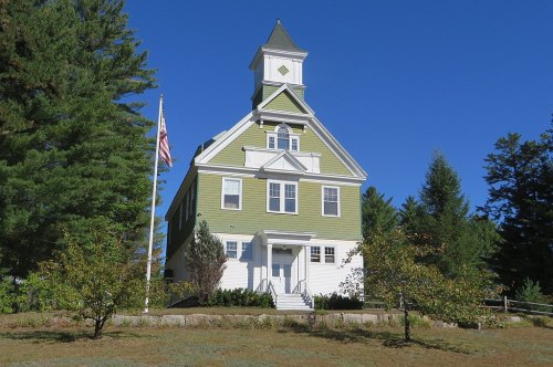 A green, three-story building with a tower, surrounded by trees and a flagpole, under a clear blue sky.
