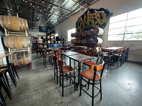 Interior of a brewery with wooden tables, chairs, and barrels stacked against the wall, featuring large windows.
