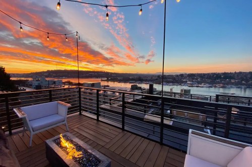 A rooftop view at sunset, featuring a fire pit, string lights, and colorful clouds over a waterfront cityscape.
