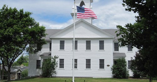 A white building with a flagpole displaying the American flag, surrounded by green trees and a cloudy sky.