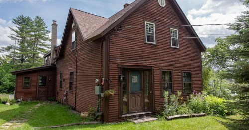 A rustic brown house with a sloped roof, surrounded by greenery and flowers on a sunny day.