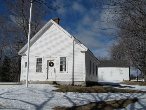 A white, historic building with a wreath on the door, surrounded by snow and bare trees under a cloudy sky.