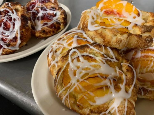 A plate of flaky pastries topped with peach slices and drizzled with icing, alongside a plate of berry-filled pastries.