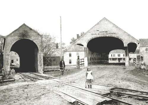 Historic black-and-white photo of a railroad crossing with a child and an adult near the tracks.