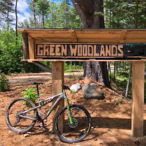 A mountain bike parked beside a wooden sign that reads "Green Woodlands" in a forested area.