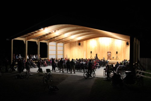 A large audience gathers under a wooden pavilion at night, watching a performance on stage. Bicycles are parked nearby.