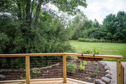 A wooden deck overlooks a lush green yard with trees and shrubs in the background under a cloudy sky.
