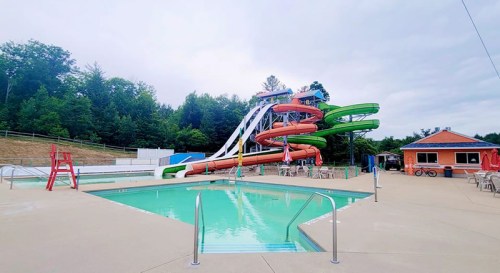 A water park featuring a large pool, colorful water slides, and lounge chairs under a cloudy sky.
