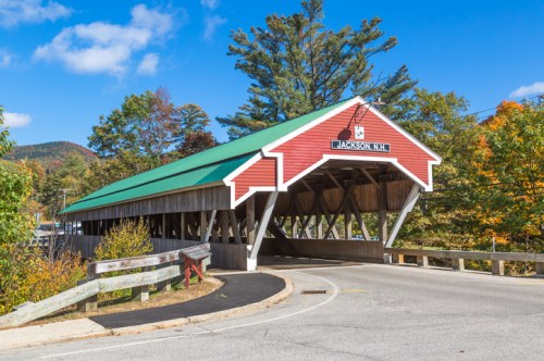 A red covered bridge with a green roof in Jackson, NH, surrounded by trees and a clear blue sky.