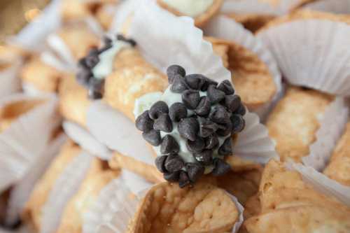 A close-up of cannoli filled with cream and topped with chocolate chips, arranged in paper liners.