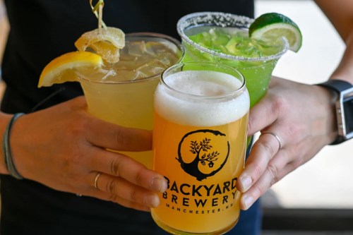 A person holds three colorful drinks: a lemon garnish cocktail, a green margarita, and a pint of beer from Backyard Brewery.