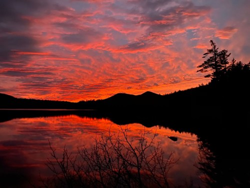 A vibrant sunset paints the sky in shades of orange and pink, reflecting on a calm lake surrounded by silhouettes of trees and hills.