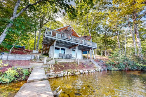 A lakeside cabin surrounded by trees, featuring a deck and a wooden walkway leading to the water.