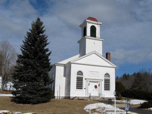 A white church with a red roof and bell tower, surrounded by snow and trees under a cloudy sky.