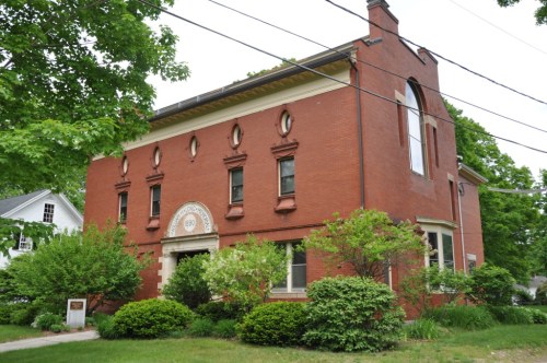 A red brick building with decorative windows, surrounded by greenery and trees, on a residential street.