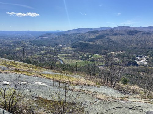 A panoramic view from a rocky hillside, showcasing rolling hills, valleys, and distant wind turbines under a clear blue sky.