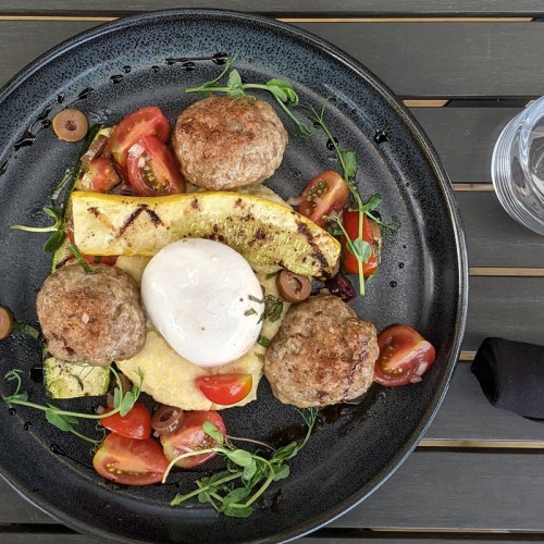 A plate with grilled zucchini, meatballs, cherry tomatoes, and a creamy burrata cheese, garnished with herbs.