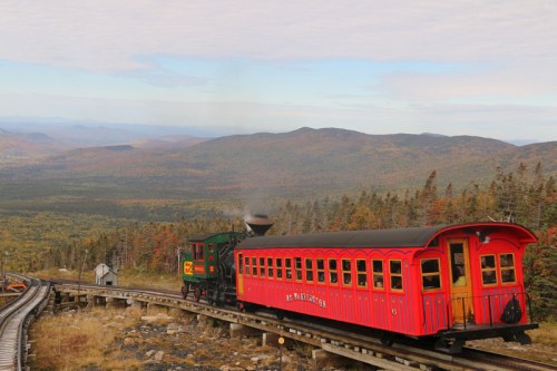 A vintage steam train travels along a winding track through colorful autumn foliage and rolling hills.