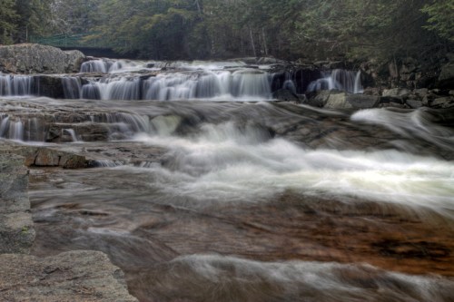 A serene waterfall cascades over rocky ledges, surrounded by lush greenery and a tranquil river.