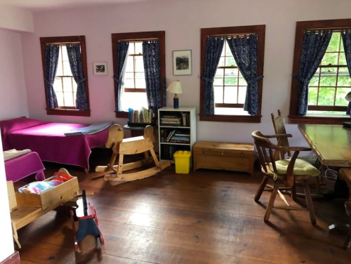 A cozy room with a bed, rocking horse, bookshelves, and a wooden table, featuring purple walls and large windows.