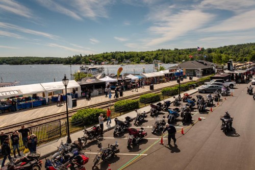 A scenic waterfront view with parked motorcycles, vendors, and a clear blue sky over a lake surrounded by greenery.