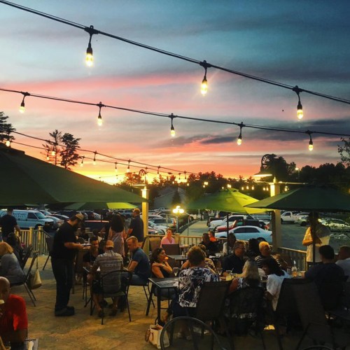 Outdoor dining scene at sunset, with string lights and patrons enjoying meals under green umbrellas.