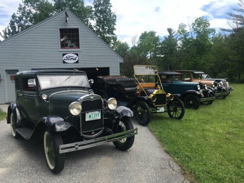 A row of vintage cars parked in front of a garage with a Ford sign, surrounded by greenery and a cloudy sky.