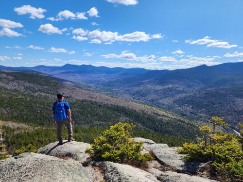A hiker stands on a rocky outcrop, overlooking a vast valley and mountains under a blue sky with scattered clouds.