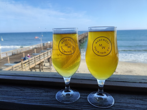 Two glasses of beer sit on a wooden table, overlooking a beach and pier with ocean waves in the background.
