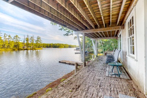 A serene lakeside view from a wooden porch with chairs, surrounded by trees and calm water under a clear sky.