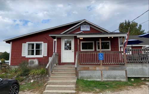 A red wooden building with a porch, sign, and accessible entrance, surrounded by greenery and a cloudy sky.