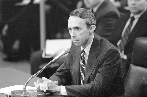 A man in a suit sits at a microphone, testifying before a committee, with a glass of water nearby.
