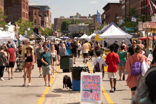 A busy street fair with crowds, tents, and vendors, featuring people walking and a sign that says "Free Water."