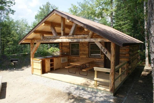 A rustic log cabin with a porch, picnic table, and outdoor kitchen, surrounded by trees and a clear blue sky.
