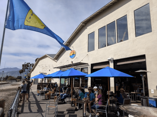 Outdoor seating area of a restaurant with blue umbrellas, people dining, and a flag waving in the breeze.