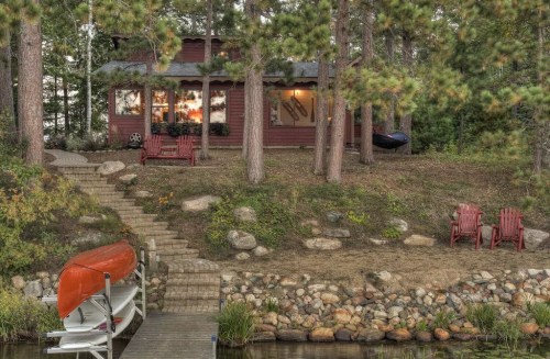 A cozy cabin by a lake, surrounded by trees, with a wooden dock and red canoe. Two red chairs sit on the shore.
