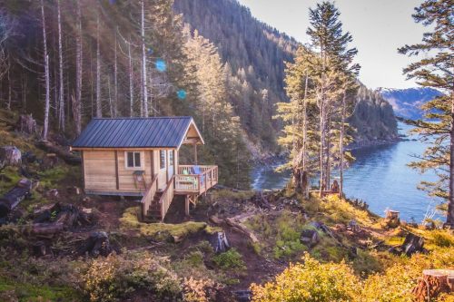A small wooden cabin by a serene lake, surrounded by tall trees and a scenic landscape.