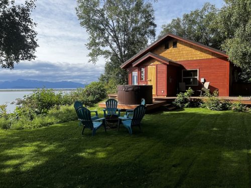 A cozy red cabin by the water, surrounded by greenery, with a hot tub and blue chairs on the lawn.